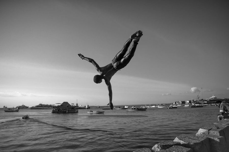 Diver, Zanzibar, Tanzania: black and white photograph of a man diving in a one-handed handstand off a harbour wall. Photograph by Marcus Vallance.